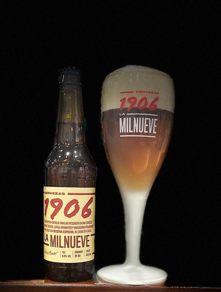 Botella de cerveza 1906 Reserva Especial junto a un vaso con cerveza servida y espuma, con iluminación focalizada.