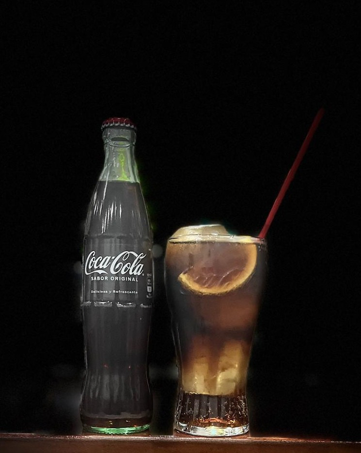 Botella de Coca-Cola con un vaso servido con hielo y rodaja de limón, sobre fondo negro.