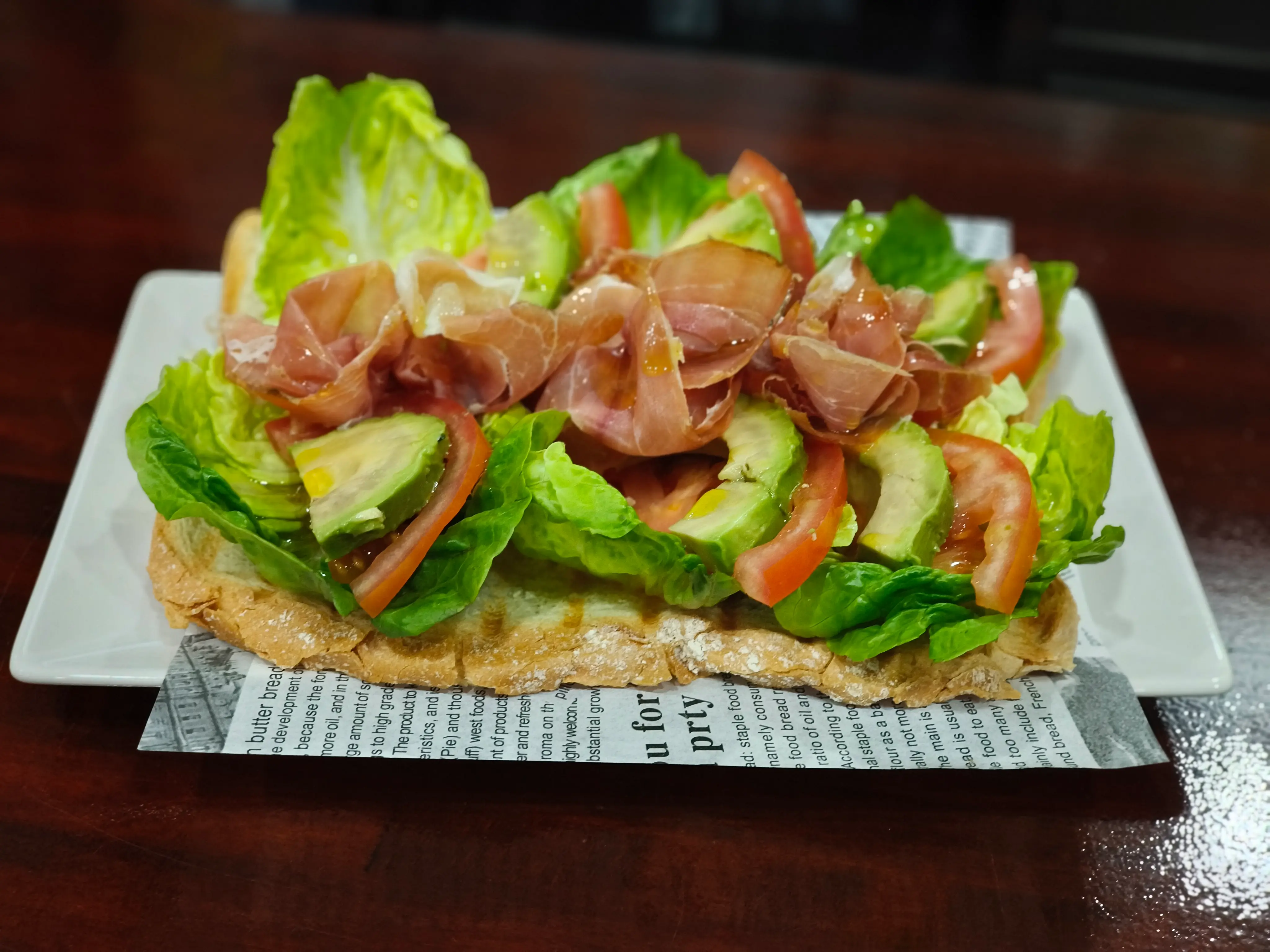 Torrada de tomate, rúcula, aguacate y jamón serrano, servida sobre pan artesanal y presentada en un plato blanco.