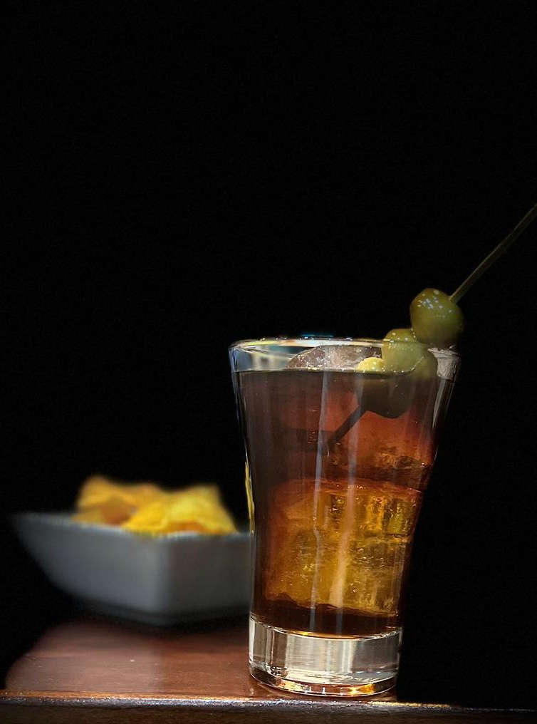 Vaso de vermut con hielo y olivas, acompañado de un plato de patatas al fondo, sobre fondo oscuro.