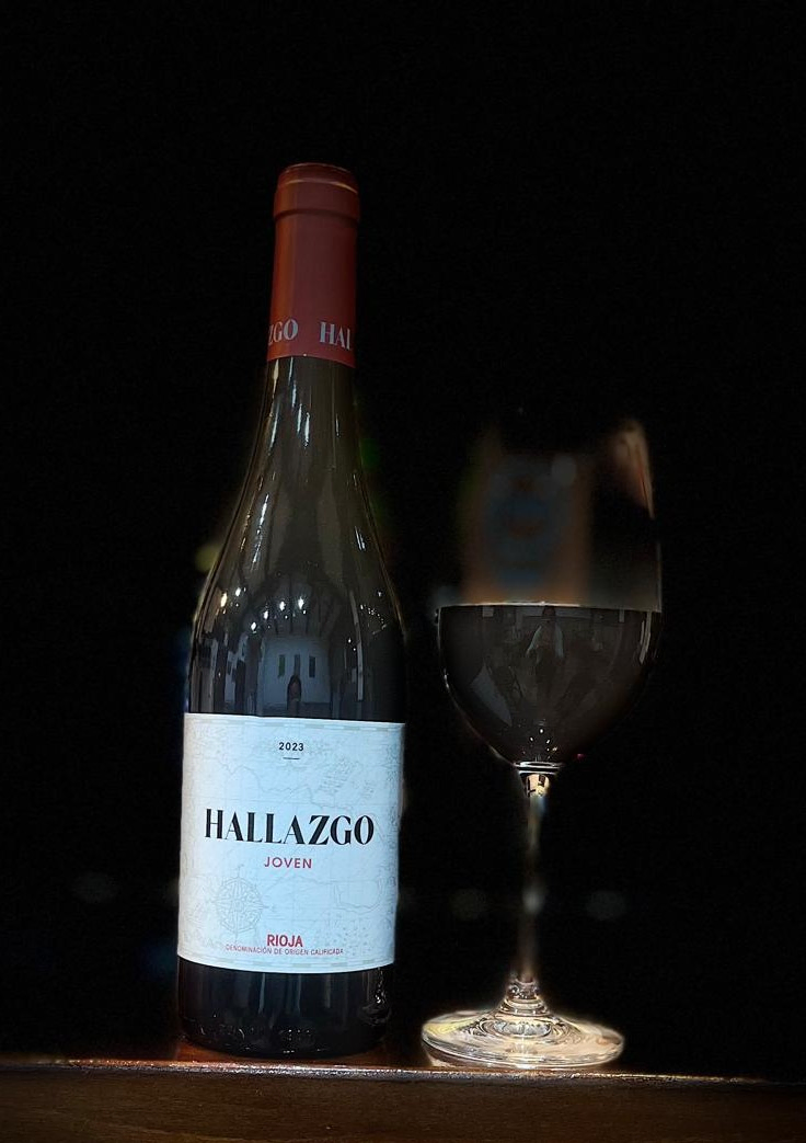 Copa de vino tinto servida y botella de vino Hallazgo, ambas con luz focalizada sobre fondo oscuro.